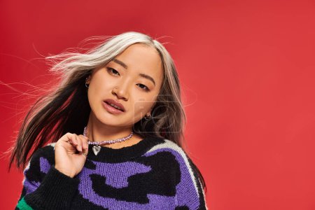 beautiful young asian woman in vibrant sweater with animal print touching necklace on red backdrop