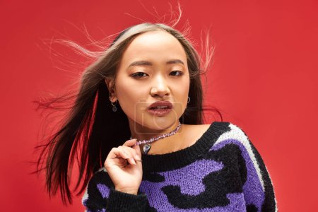 pretty young asian woman in vibrant sweater with animal print touching necklace on red backdrop
