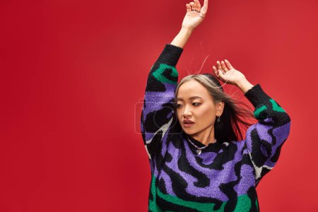 alluring young asian woman in vibrant sweater with animal print posing with raised hands on red