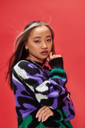 Foto de Hermosa joven asiático modelo en vibrante suéter con animal print tocando mejilla en rojo telón de fondo - Imagen libre de derechos
