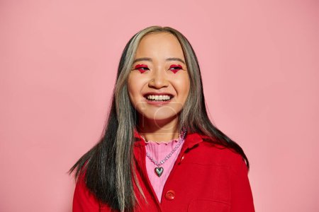 San Valentín concepto de día, alegre asiático mujer con corazón en forma de ojo maquillaje posando sobre rosa telón de fondo