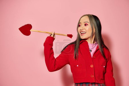 Valentines day concept, jolly asian woman with heart shaped eye makeup holding arrow on pink