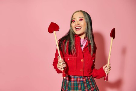 Valentines day, excited asian cupid girl with heart shaped eye makeup holding arrows on pink