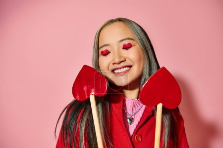 Valentines day concept, jolly asian girl with heart shaped eye makeup holding arrows on pink