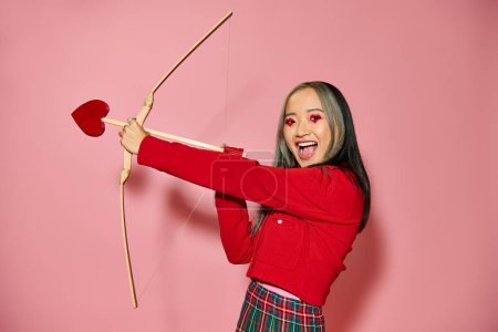 Valentines day, jolly asian woman with heart shaped eye makeup holding cupid arrow and bow on pink