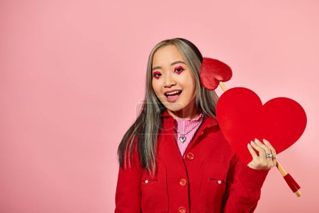 Valentines day, excited asian woman with vibrant eye makeup holding carton heart on pink backdrop