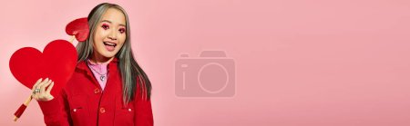 Valentines day banner, excited asian woman with eye makeup holding carton heart on pink backdrop