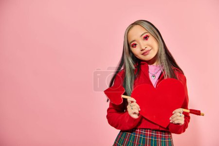 Valentines day, smiling asian woman with vibrant eye makeup holding carton heart on pink backdrop