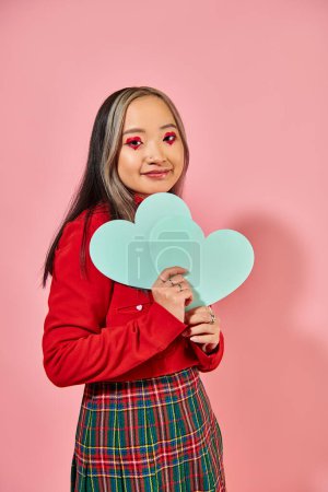 Valentines day, happy asian girl with vibrant eye makeup holding blue carton hearts on pink backdrop