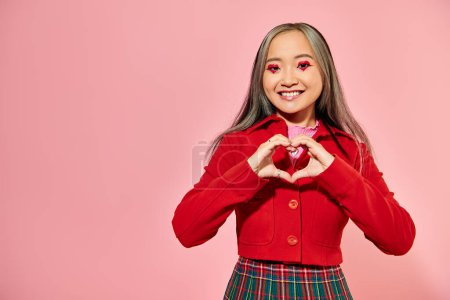 Día de San Valentín, feliz chica asiática con maquillaje de ojos rojos mostrando el corazón con las manos en el fondo rosa