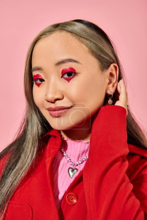 Valentines day, positive asian young woman with heart eye makeup posing in red jacket on pink