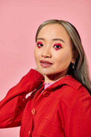 Valentines day concept, pretty asian young woman with heart eye makeup posing in red jacket on pink