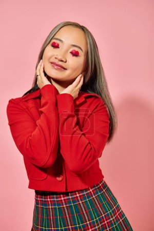 Valentines day, pleased asian young woman with heart eye makeup posing in red jacket on pink