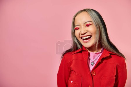 San Valentín concepto de día, feliz mujer asiática con el corazón maquillaje de ojos riendo sobre fondo rosa