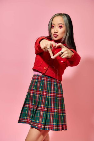 Valentines day concept, young asian girl in red jacket showing heart with fingers on pink backdrop