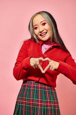 San Valentín concepto de día, feliz asiática chica en rojo chaqueta mostrando corazón con los dedos en rosa telón de fondo