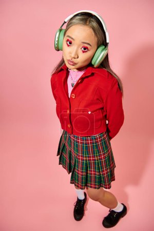 Foto de San Valentín maquillaje día, joven mujer asiática en verde auriculares inalámbricos posando en rosa telón de fondo - Imagen libre de derechos