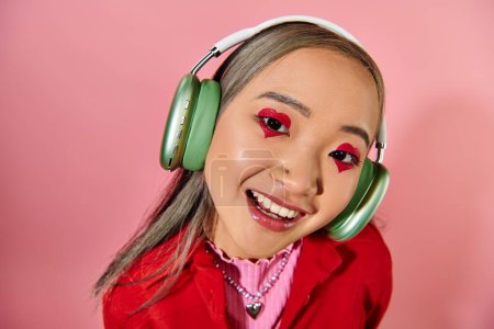 happy young asian woman in green wireless headphones with heart shaped makeup posing on pink