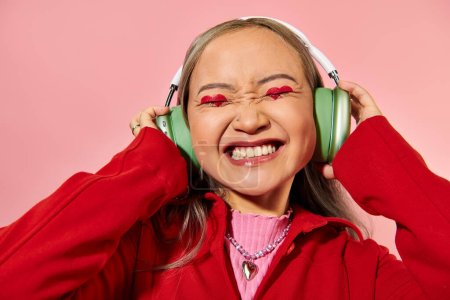 joyful young asian woman in green wireless headphones with heart shaped makeup on pink backdrop