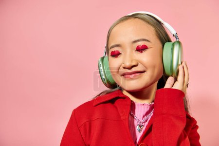jolly young asian woman in green wireless headphones with heart shaped makeup on pink backdrop