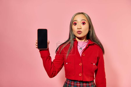 young asian woman with red Valentines day makeup showing smartphone with blank screen on pink
