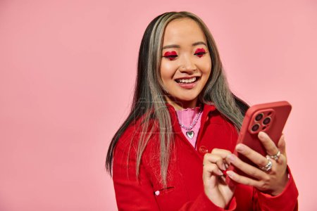 cheerful and young asian woman with red Valentines day makeup using smartphone on pink backdrop