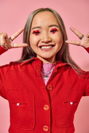 happy asian young woman in red jacket showing v sign on pink background, heart shape eye makeup
