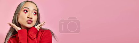 Foto de Bonita asiática joven mujer con corazón forma ojo maquillaje puting labios y mirando hacia otro lado en rosa, bandera - Imagen libre de derechos