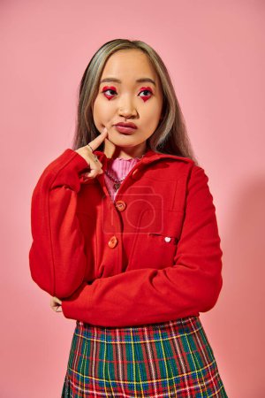 Foto de Pensativo asiático joven mujer con corazón forma ojo maquillaje tocando mejilla y mirando lejos en rosa - Imagen libre de derechos