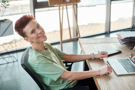 stylish happy non-binary person looking at camera white sitting near laptop at workplace in office