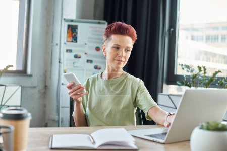 Téléchargez les photos : Élégant bigender individu avec smartphone travaillant à l'ordinateur portable près du portable dans le bureau moderne - en image libre de droit