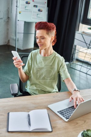 smiling bigender individual looking at smartphone near laptop and notebook in modern office