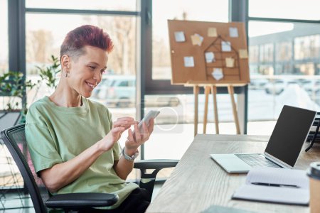Lächelnde bigeschlechtliche Person mit Smartphone in der Nähe von Laptop und Notebook am Arbeitsplatz im modernen Büro