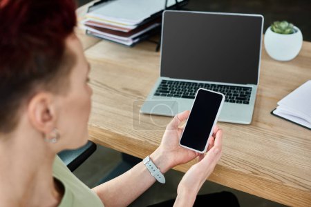 Téléchargez les photos : Personne d'affaires non binaire tenant smartphone avec écran vierge près d'un ordinateur portable au travail dans le bureau - en image libre de droit