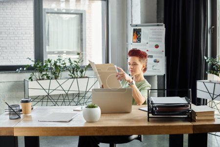 stylish bigender manager working with documents near laptop and coffee to go in paper cup in office