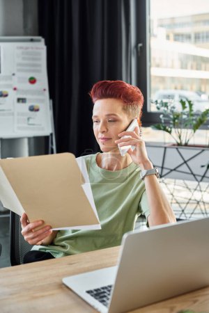 persona queer ocupado hablando en el teléfono inteligente y mirando los documentos cerca de la computadora portátil en la oficina moderna