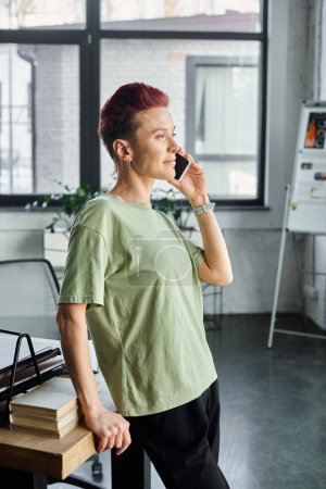 stylish non-binary manager in casual attire talking on smartphone while standing in modern office