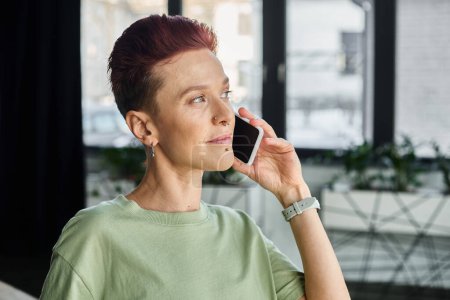 Stilvoller Non-binary-Manager in lässiger Kleidung, der im modernen Büro mit dem Smartphone spricht