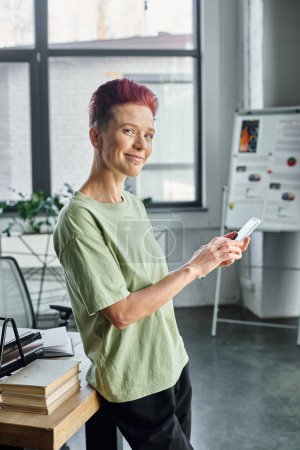 cheerful successful queer manager with mobile phone looking at camera while standing in office