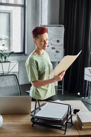 stylish queer manager holding folder with documents and standing near laptop on work desk in office