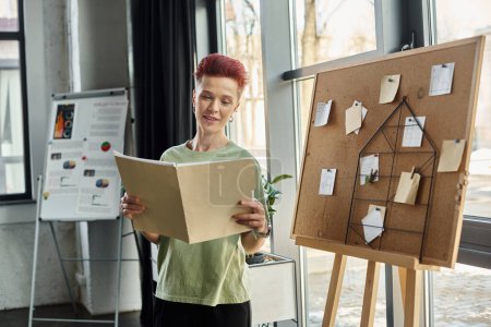 smiling queer person looking at folder with documents near corkboard with paper notes in office