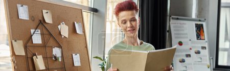 smiling queer person looking at documents near corkboard with paper notes in office, banner