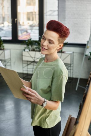 positive non-binary manager with short hair looking at folder with documents while working in office