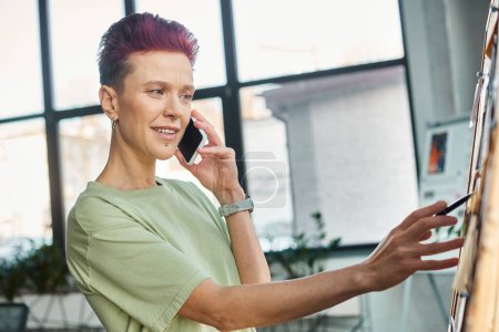 Stilvolle queere Person, die mit dem Handy telefoniert und im Büro Zettel auf Korkpappe betrachtet