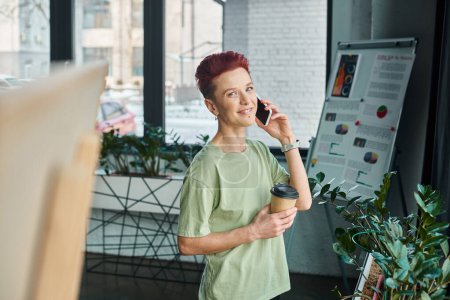 glückliche queere Person mit Coffee to go in Pappbecher im Gespräch mit dem Handy im modernen Büro