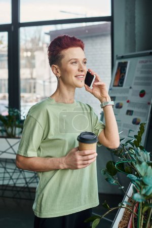 feliz persona queer con café para ir en taza de papel hablando en el teléfono móvil en la oficina moderna