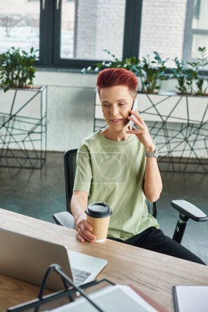 persona queer positivo con café para ir en taza de papel hablando en el teléfono móvil cerca de la computadora portátil en la oficina