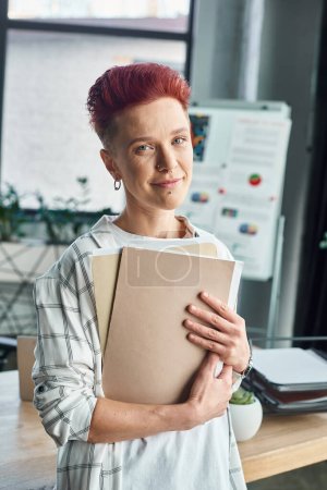 portrait of non-binary manager holding folders with documents and looking at camera in office