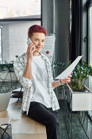 queer person in casual attire sitting on work desk with document and talking on smartphone in office