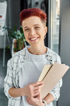 portrait of cheerful queer person holding folder with documents and looking at camera in office